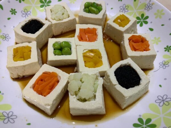 將切成正方塊的豆腐中間挖空，分別放入代表白色的白花椰菜、代表黃色的甜椒、代表綠色的毛豆、代表橘紅色的胡蘿蔔和代表黑色的髮菜，搭配醬汁燴煮。