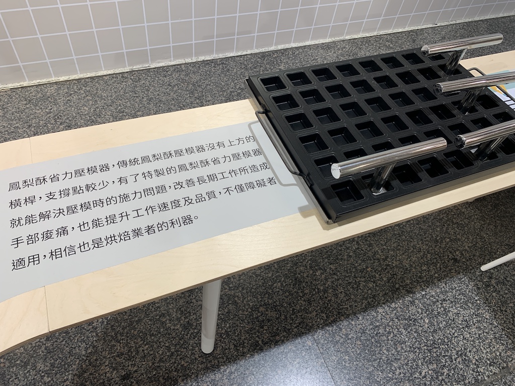 畫面中右邊是鳳梨酥省力壓模器，黑色的壓模器上面有一個個方形的洞，跟鳳梨酥的大小一樣。上面還有數個壓模的金屬握桿。畫面左邊是一段介紹文字：「鳳梨酥省力壓模器，傳統鳳梨酥壓模器沒有上方的橫桿，支撐點較少，有了特製的鳳梨酥省力壓模器，就能解決壓模時的施力問題，改善長期工作所造成手部痠痛，也能提升工作速度及品質，不僅障礙者適用，相信也是烘焙業者的利器。