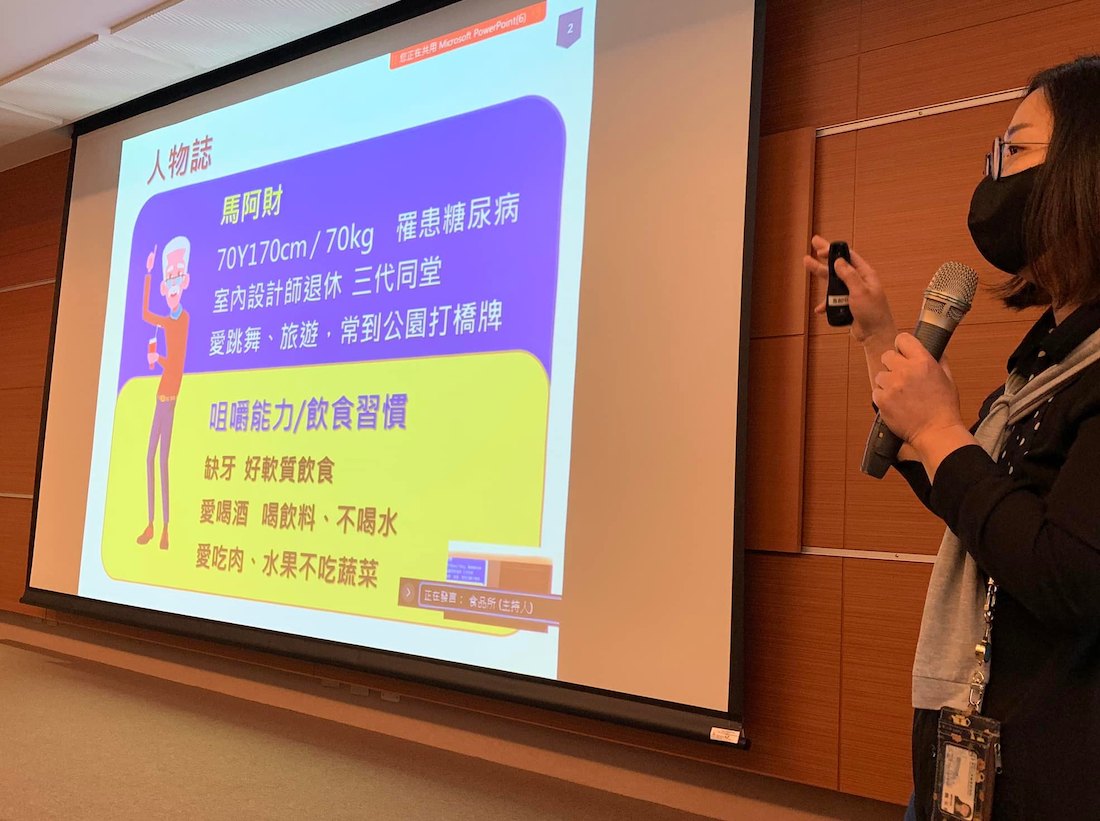 照片中主要是一個投影幕,投影幕上面有簡報畫面,右邊站著一個拿著麥克風和簡報器,並戴著口罩在說話的人。